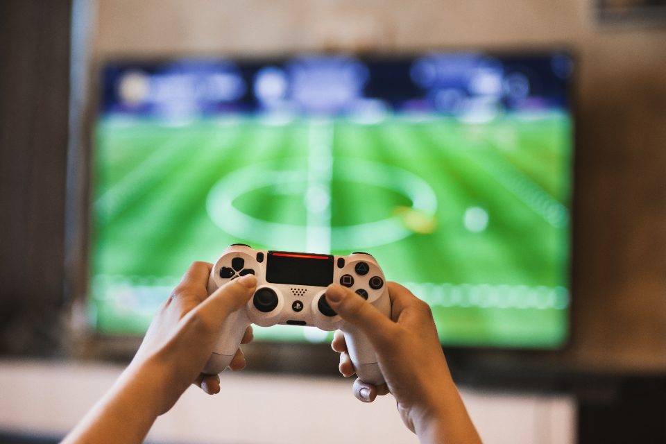 To hænder holder en hvid PS4-controller foran et fjernsyn med et fodboldspil