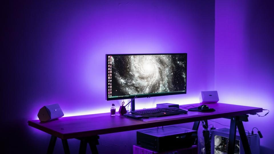 En bred pc-skærm med tastatur og mus på et skrivebord i lilla LED-lys